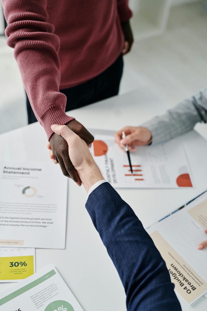Professional handshake across a desk with financial charts and business reports